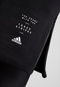 Tissu noir avec texte blanc "THE BRAND WITH THE THREE STRIPES" et logo Adidas au-dessus d'une fermeture éclair partiellement visible.