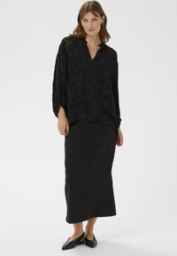 Juoda tekstūruota bliuska su V formos iškirpte ir varlės rankovėmis, dėvima su ilga juoda sijonu. Modelis vilki juodus aštrius batus.