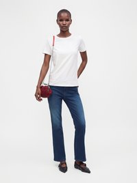 Femme portant un t-shirt blanc, un jean bleu, des chaussures pointues noires, et portant un petit sac rond rouge avec une bandoulière en chaîne.