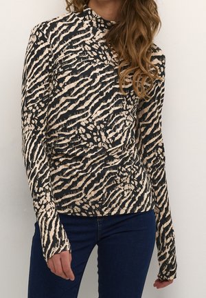 Frau trägt ein langärmliges, hochgeschlossenes Oberteil mit beigem und schwarzem Tierdruck, kombiniert mit dunkelblauen Jeans vor weißem Hintergrund.