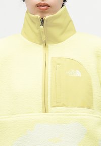 Άτομο που φοράει ανοιχτό κίτρινο φλις μπουφάν με φερμουάρ στον ψηλό γιακά και το λογότυπο The North Face στην αριστερή τσέπη στο στήθος.