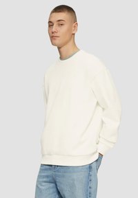 Biała bluza z okrągłym dekoltem, długimi rękawami i prążkowanymi mankietami. Noszona z jasnoniebieskimi dżinsami o gładkiej fakturze.