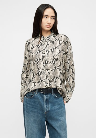 Woman with long dark hair wearing a long-sleeve snake print blouse tucked into loose blue jeans with a black belt.