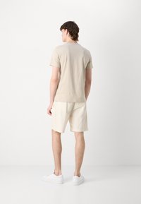 Beige pamut póló rövid ujjakkal, világos krém színű sorttal párosítva. A lábbelik fehér sportcipők. A háttér egyszínű fehér.