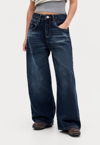 Woman wearing dark blue wide-leg jeans with hands in pockets, paired with brown shoes and a cropped striped top.