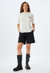 Camiseta de algodón blanca de manga corta con detalle bordado negro, combinada con pantalones cortos negros y botas de cuero negras hasta la rodilla.