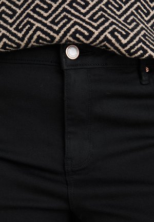 Mustade pükste lähedalähedane vaade, millel on hõbedane nööp ja taljesse toppinud beežide ja mustade geomeetrilise mustriga kangas.