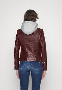 Giacca in pelle color mogano con design aderente, dettagli di cuciture sottili e cappuccio grigio. Indossata con jeans blu.