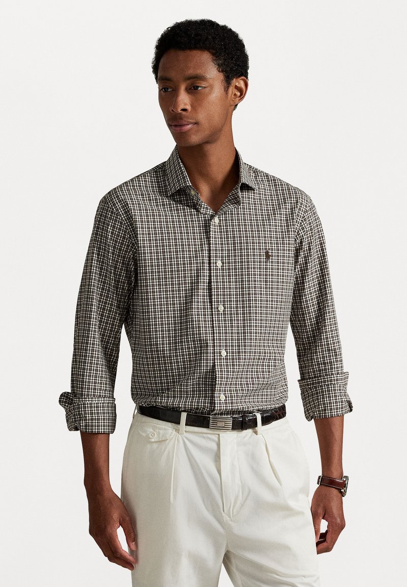 Chemise à carreaux brun et blanc avec un col pointu, des manches roulées et un petit logo sur la poitrine. Portée avec un pantalon de couleur claire.