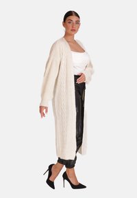 Długi, dziany kardigan w kolorze kremowym z wzorem w warkocze, zestawiony z czarnymi dopasowanymi spodniami i czarnymi butami na obcasie. Gładka tekstura.