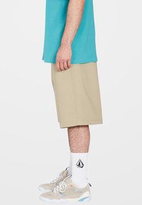 Beige shorts met een losse pasvorm, gecombineerd met een blauwe t-shirt. De schoenen zijn beige met lichte blauwe accenten, gedragen met witte sokken met een logo.