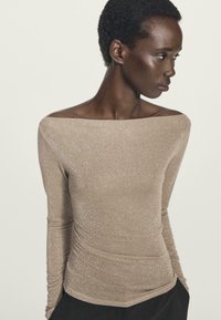 Donna con capelli ricci corti che indossa un top lungo a maniche a sbuffo beige glitterato, che guarda verso destra su uno sfondo neutro.