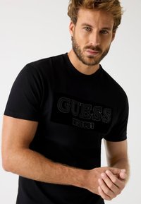 Czarny bawełniany t-shirt z wyraźnym czarnym logo "GUESS" oraz tekstem "EST. 1981" w prostokącie. Krótkie rękawy, okrągły dekolt.