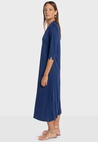 Marineblauwe jurk tot op de knie met driekwartmouwen, voorzien van een gespleten detail bij de manchetten en een gladde textuur. Draagt bijpassende sandalen met bandjes.