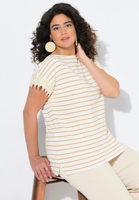 Pruhovaný top v krémové a žluté barvě, s krátkými rukávy lemovanými krajkou. Boční rozparky a uvolněný střih, v kombinaci s béžovými kalhotami.