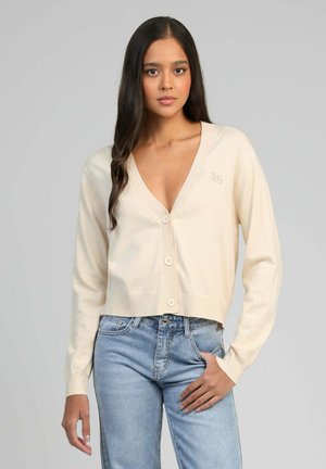 Jeune femme aux longs cheveux bruns portant un cardigan crème à col en V boutonné et un jean bleu clair, posant avec une main dans la poche.