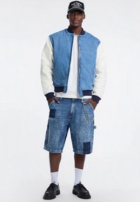 Gaiši zils un krēmkrāsas džinsa bomber stila jaka ar ribveida aprocēm, saskaņota ar bojātiem zilajiem šortiem ar plāksteriem un ķēdes detaļu.