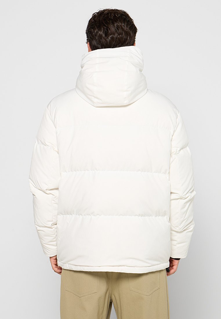 Giacca puffer bianca con cappuccio, caratterizzata da un design trapuntato e cuciture orizzontali. Tessuto morbido e vestibilità rilassata. Vista posteriore.