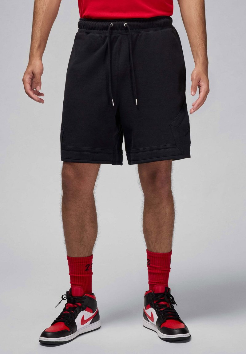 Pantalones cortos de forro polar negros con cintura elástica y cordón. Presentan bolsillos laterales y un detalle de patrón cosido. Combinados con calcetines rojos y zapatillas negras/rojas.