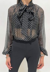 Donna che indossa una blusa nera trasparente con pois bianchi e un grande fiocco, abbinata a pantaloni neri a vita alta e braccialetti.
