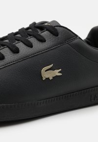 Černá kožená teniska s texturovaným povrchem, černými tkaničkami a zlatým logem krokodýla na boku, s gumovou podrážkou s geometrickými vzory.