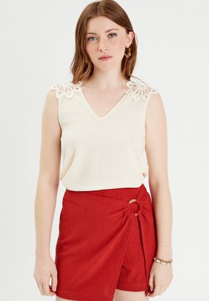 Top crema con scollo a V e dettaglio in pizzo sulle spalle, abbinato a pantaloni corti rossi dalla texture con design avvolgente e accentuazione in metallo circolare.