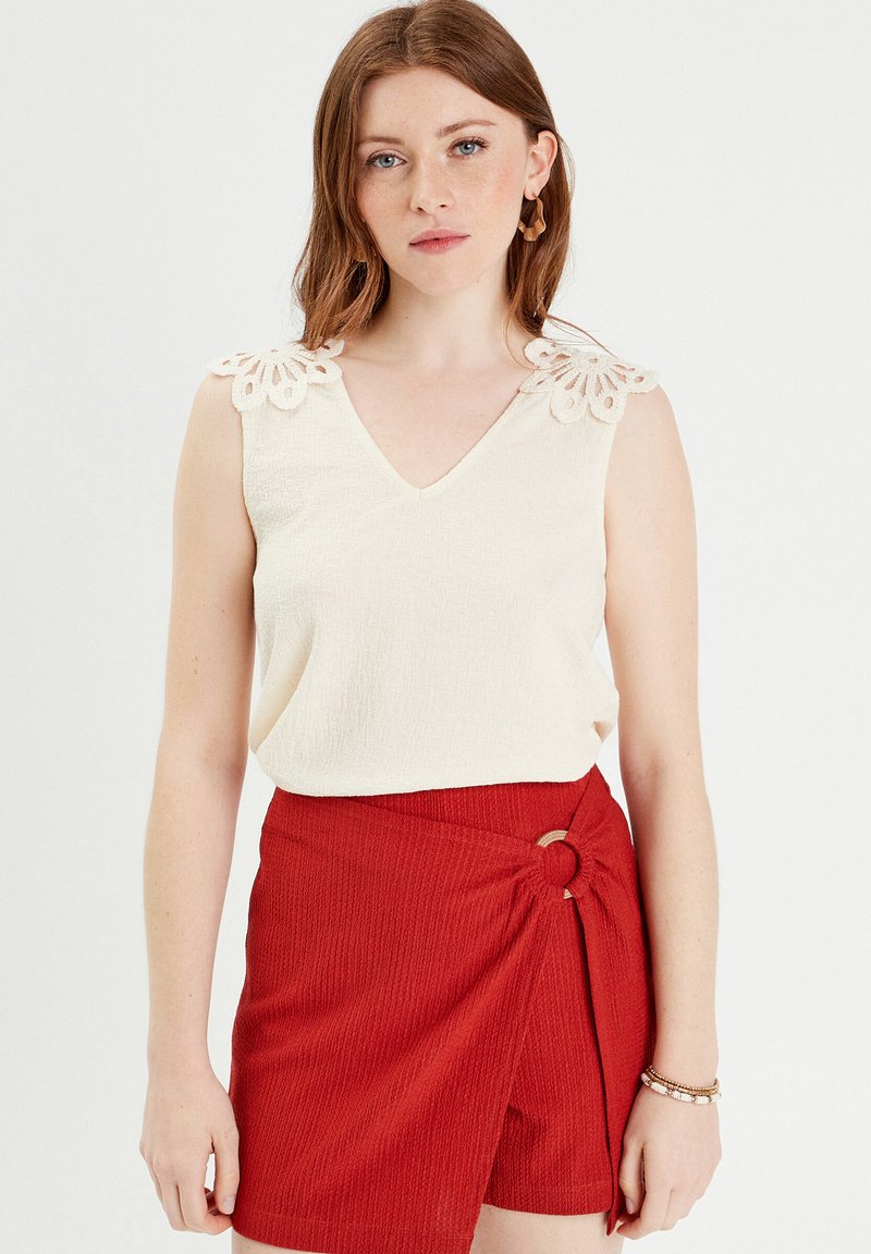 Haut crème en v avec des détails en dentelle aux épaules, associé à un short rouge texturé avec un design enrobé et un accent en métal circulaire.