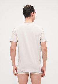 Mann, der mit dem Rücken zur Kamera steht und ein schlichtes, hellbeiges kurzärmliges Hemd sowie passende Shorts trägt, vor einem schlichten weißen Hintergrund.