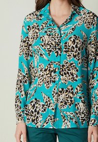 Woman wearing turquoise button-up blouse with large beige and black leopard print leaves, paired with dark green pants.