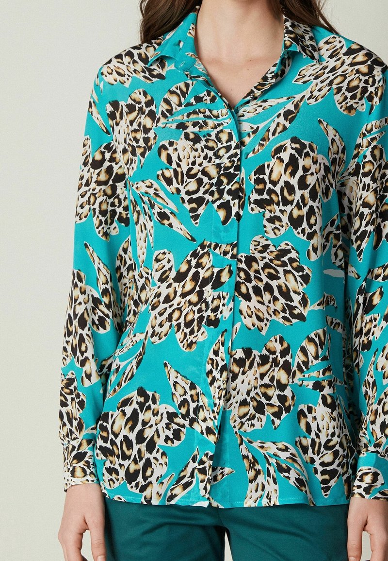 Woman wearing turquoise button-up blouse with large beige and black leopard print leaves, paired with dark green pants.