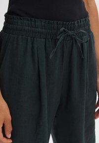 Donkergroene linnen broek met een elastische tailleband, verstelbaar trekkkoord en zijplooien voor een ontspannen pasvorm. Zachte textuur, casual ontwerp.