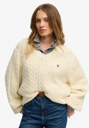 Pull en laine crème à tissage cablé avec un col en V, doté de manches amples et d'un petit logo. Porté par-dessus une chemise à col rayée bleu et blanche.