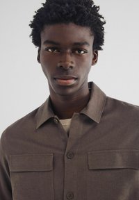 Bruine button-up shirt met twee borstzakken, een textuurstof en een klassieke kraag, in laagjes over een lichte trui.