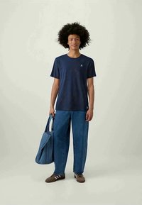 Young person with curly hair wearing a navy blue t-shirt, loose blue jeans, brown sneakers, holding a large denim tote bag.