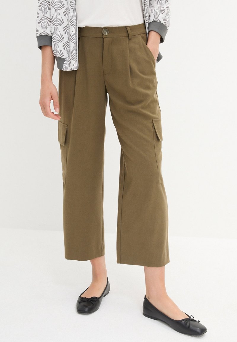 Personne portant un pantalon cargo court olive avec poches latérales, des ballerines noires, un haut blanc et une veste légère à motifs sur un fond uni.
