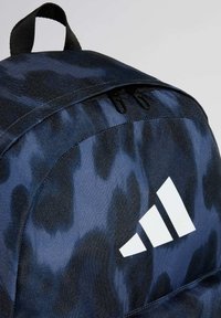 Plava ruksak s teksturiranim materijalom koji prikazuje tamni apstraktni uzorak, s gornjom ručkom i istaknutim bijelim Adidas logotipom na prednjoj strani.