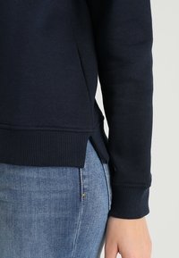 Felpa blu navy con polsini e orlo a costine, caratterizzata da fessure laterali per un tocco di dettaglio. Indossata con jeans in denim azzurro.