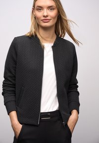 Zwarte bomberjack met ruitstiksel en visgraatpatroon, geribde kraag, ritssluiting en zijzakken, gedragen over een wit shirt en zwarte broek.