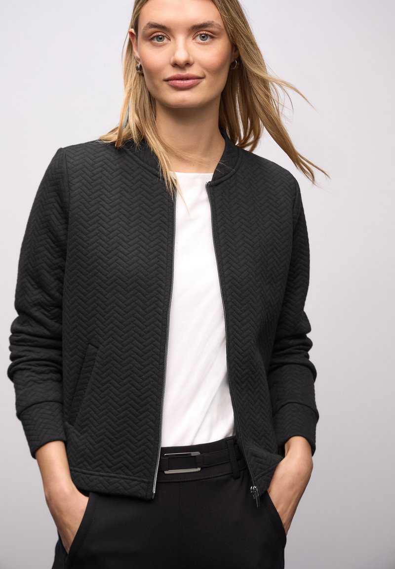 Zwarte bomberjack met ruitstiksel en visgraatpatroon, geribde kraag, ritssluiting en zijzakken, gedragen over een wit shirt en zwarte broek.