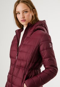 Bordo puffer jakna s kapuljačom, koja ima kvadratni dizajn, prednji zip, elastičan struk i detalj s logotipom na rukavu. Glatka tekstura.