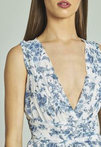 Vestido floral en blanco con patrones azules. Presenta un escote en V profundo, diseño sin mangas y cintura fruncida para una silueta ajustada.