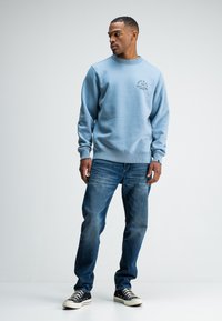  Lichtblauwe sweatshirt met geribde boorden en onderkant, voorzien van een geborduurd logo. Gecombineerd met vervaagde blauwe spijkerbroek en zwarte canvas sneakers.