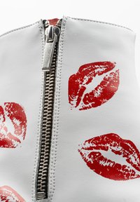 Botín de tobillo de imitación cuero blanco con una cremallera plateada y estampados de labios rojos en la superficie, con una textura suave y forma redondeada.