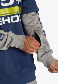 Marinblå och grå sweatshirt med en stor logotyp, som har en unik ärmficka och ribbade muddarna, tillverkad av mjukt texturerat tyg.