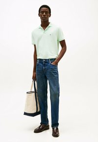 Lichtgroene polo, blauwe denim jeans, bruine leren schoenen, met een beige tote bag met marineblauwe accenten. Eenvoudig en casual ontwerp.