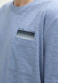 Camiseta de cuello redondo en azul claro, de tela suave, que cuenta con un pequeño bolsillo en el pecho con un estampado gráfico y el logotipo de la marca. Textura lisa, diseño casual.