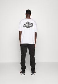 Vitt T-shirt med en svart Los Angeles Lakers-logotyp på ryggen, kombinerat med svarta sweatpants och svarta sneakers.