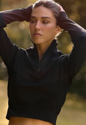 Sort, ribbet atletik top med høj krave og halv-lynlås. Den cropped design fremhæver taljen. Lange ærmer har tætsiddende manchetter.