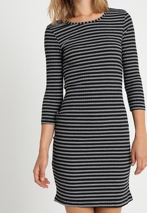 Mujer con vestido negro con rayas horizontales blancas, mangas tres cuartos y escote redondo, de pie frente a un fondo blanco liso.