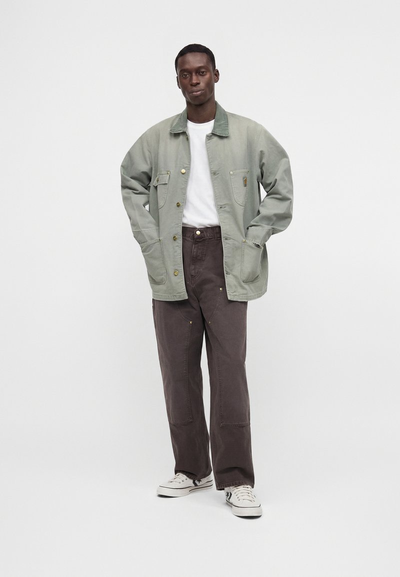 Mann steht mit den Händen in den Taschen, trägt eine hellgrüne Jacke, ein weißes T-Shirt, dunkelbraune Hose und weiße Turnschuhe vor einfarbigem Hintergrund.